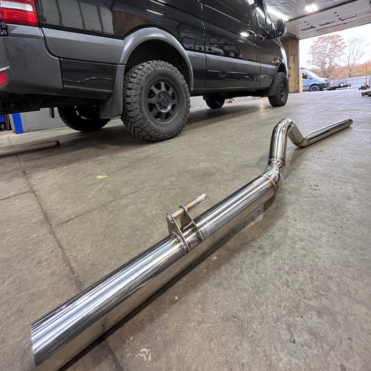 A silver stainless steel tail pipe for a Revel stainless 144" 2500 van/ Revel Stainless 304 exhaust system, displayed on a shop floor with a van in the background.