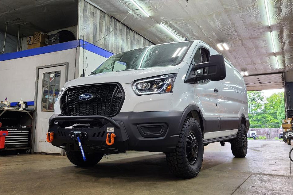 A white Ford Transit van parked inside a garage workshop, featuring a black front grille, off-road tires, a front bumper winch, and orange recovery hooks.