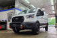 A white Ford Transit van parked inside a garage workshop, featuring a black front grille, off-road tires, a front bumper winch, and orange recovery hooks.