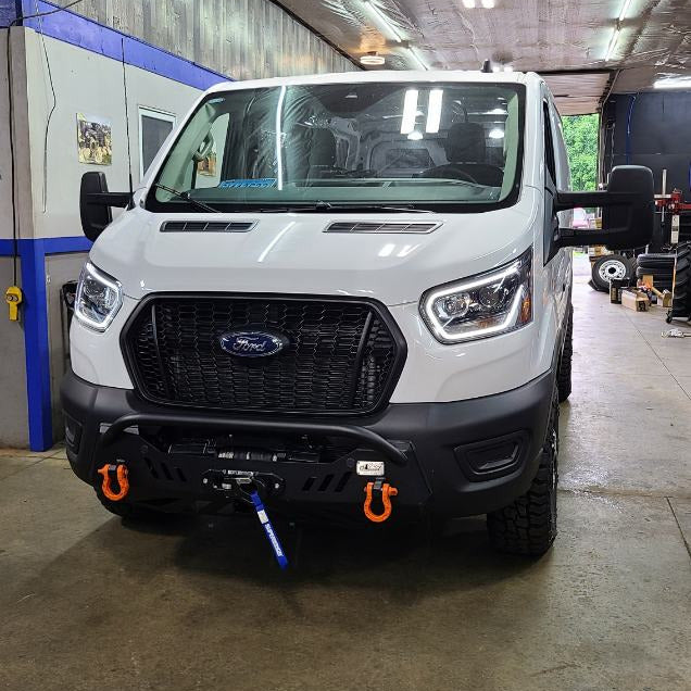 A white Ford Transit van parked inside a garage or workshop, shown from the front. The vehicle has a black front grille and bumper, with orange tow hooks on the front bumper.