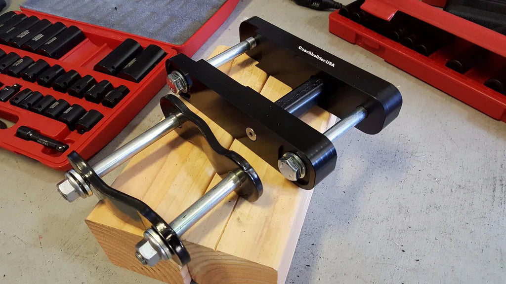A black stock Toyota shackle with silver bolts and nuts, sitting on a wooden block next to a coachbuilder extended shackle for comparison.