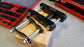 A black stock Toyota shackle with silver bolts and nuts, sitting on a wooden block next to a coachbuilder extended shackle for comparison.