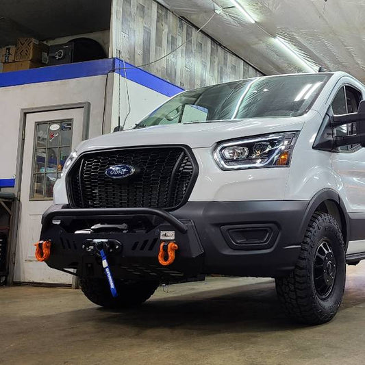 A white Ford Transit van with a black front bumper and off-road tires parked inside a workshop or garage. A person is partially visible on the right holding a camera or phone.