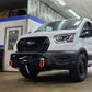 A white Ford Transit van with a black front bumper and off-road tires parked inside a workshop or garage. A person is partially visible on the right holding a camera or phone.